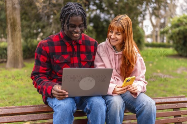 Two happy young students are using a laptop and a smartphone while sitting on a bench in a park, enjoying online learning and connectivity