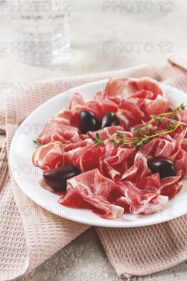 Freshly cut dried meat, served with olives and herbs, on a white plate, light background, no people