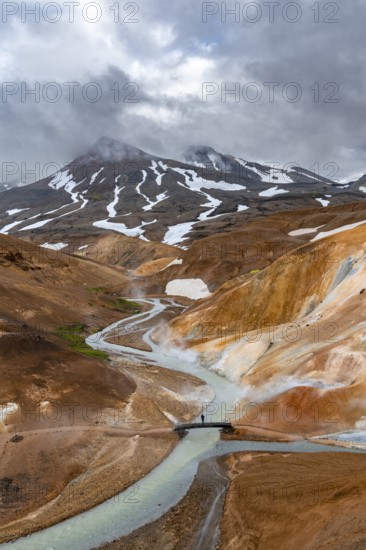 Tourist on bridge over steaming stream between colourful rhyolite mountains with snowfields, Hveradalir geothermal area, Kerlingarfjöll, Icelandic highlands, Iceland