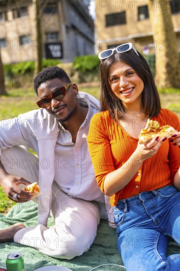 Happy interracial couple eating pizza outdoors, enjoying a relaxing picnic in a sunny urban park