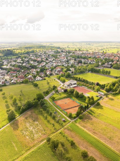 Aerial view of a town with neighbouring fields and a tennis court, surrounded by green vegetation and under a cloudy sky, Kieselbronn, Germany