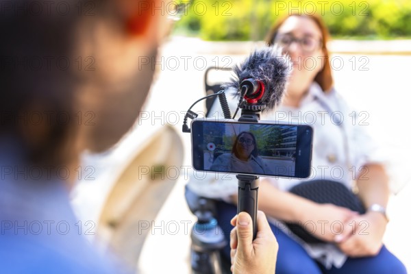Videographer holding a smartphone on a tripod with external microphone, recording an interview with a businesswoman in a wheelchair outdoors