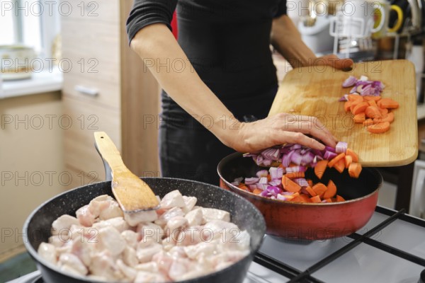 Unrecognizable woman cooking chicken pieces in one pan while pouring chopped onions and carrots to another. Natural light fills the space, enhancing the inviting atmosphere