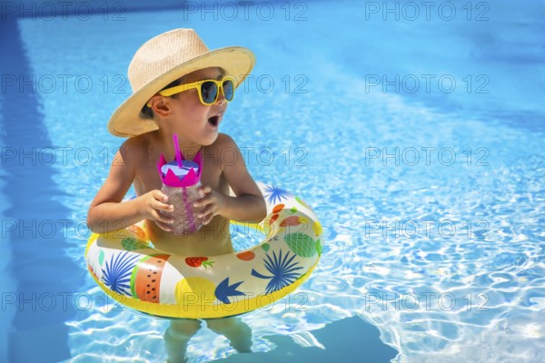 Playful caucasian male kid in sun hat and sunglasses on a pool with a glass of water standing inside a rubber ring float