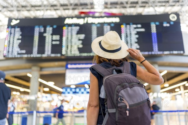 A woman wearing a straw hat and a backpack is looking at a large airport screen. She is waiting for her flight