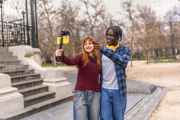 Two young content creators are recording a video for their vlog using a smartphone with a microphone attached, while standing in a park
