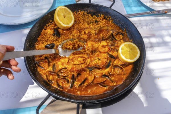 Close up of a spoon scooping up seafood paella from a traditional paella pan, garnished with lemon wedges, during a summer meal
