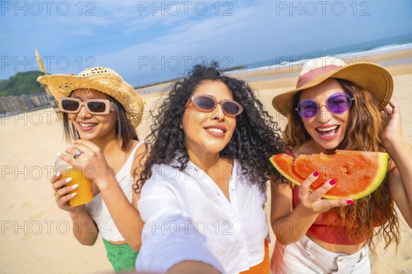 Three women are smiling and holding watermelons on a beach. They are wearing straw hats and sunglasses