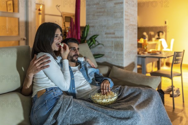 Young couple enjoying a movie night at home, eating popcorn from a bowl and relaxing on the sofa under a blanket