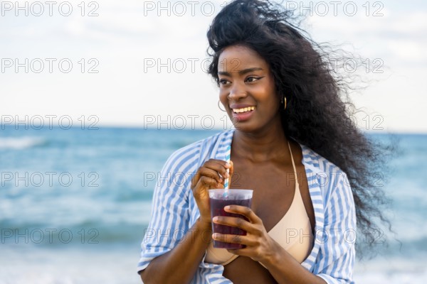 Smiling african american woman enjoying a refreshing drink on a sunny beach, with blue sea in the background