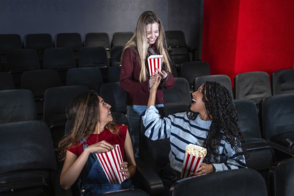 Three female friends are laughing and sharing popcorn while watching a movie in a cinema, enjoying their leisure time together