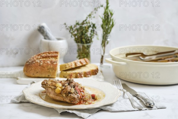 Cooked rabbit stew is served on a plate with herbs on top. Fresh bread is placed next to the dish on a simple cloth. A pot is shown nearby with more stew, creating a homey kitchen scene