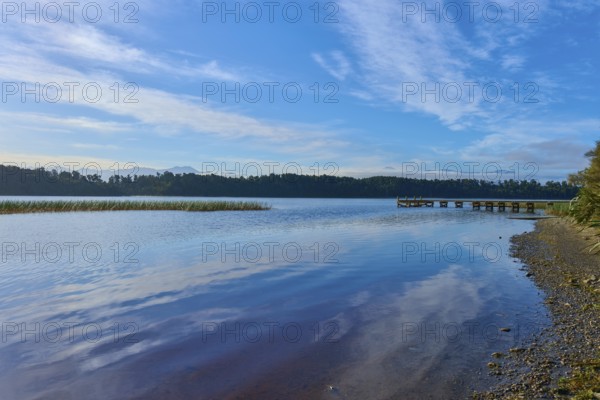 A jetty and shingle beach stretch along the shore of a tranquil lake under blue skies, Lake Mahinapua, Ruatapu, South Island, New Zealand