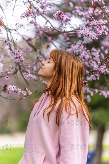 Young woman with long red hair, wearing pink hooded sweatshirt, breathing fresh air and enjoying scent of cherry blossoms in a park during springtime