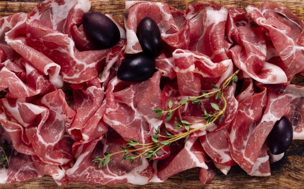 Freshly cut dried meat, served with olives and herbs, on a wooden chopping board, no people