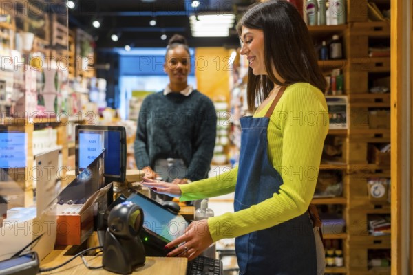 Smiling saleswoman wearing apron using touchscreen cash register while customer waiting at checkout counter in organic supermarket