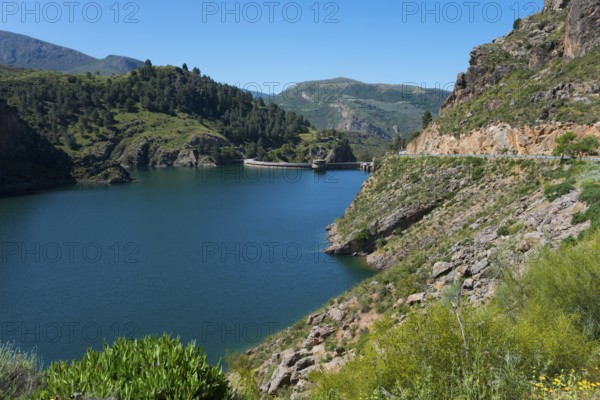 A tranquil lake in a mountainous landscape with green hills and rocks, under a clear blue sky, reservoir, Embalse de Quéntar, Quentar, Granada province, Andalusia, Spain