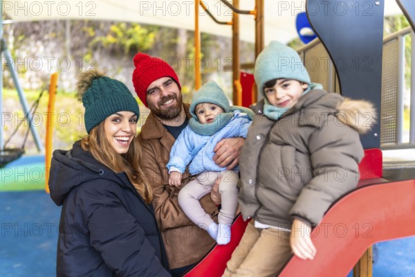 Smiling family bundled in winter clothes, enjoying quality time together while playing on a colorful playground slide in the park