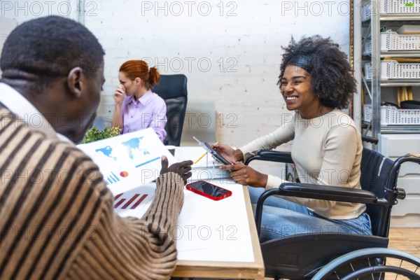Cheerful and smiling african woman with disability sitting on wheelchair and colleague discussing graphs in a coworking