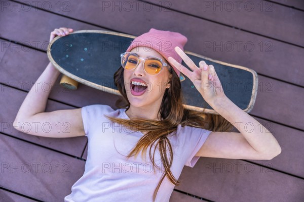 Happy young woman lying on a wooden floor with her skateboard showing peace sign, wearing pink beanie hat and yellow sunglasses, enjoying leisure activity