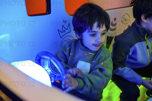 Young boy playing with toy car steering wheel, enjoying entertainment in colorful amusement park illuminated with neon lights