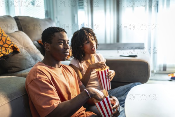 Young couple enjoying a movie night at home, sitting on the sofa, eating popcorn and watching television