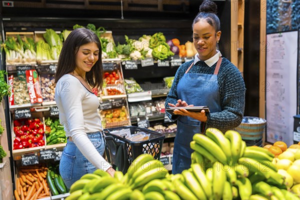 Supermarket employee using tablet helping customer buying bananas in grocery store with fresh fruits and vegetables