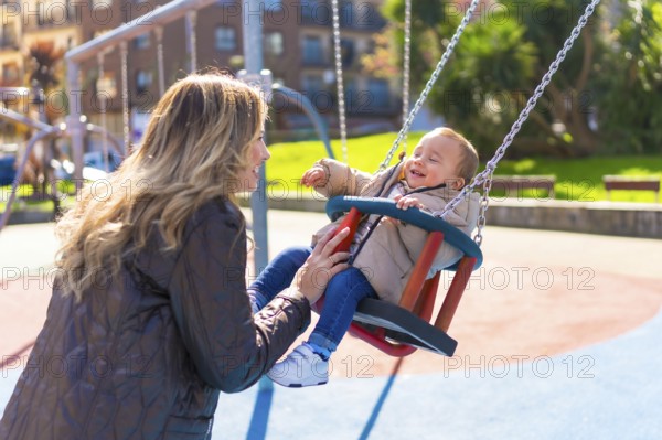 Happy mother pushing her laughing baby son on a swing, enjoying a sunny autumn day at the playground, creating joyful family memories