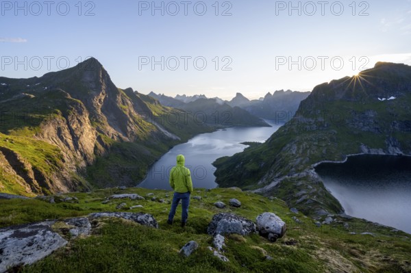 Mountaineer looking over mountain landscape with pointed mountain peaks and fjord Forsfjorden with village Vindstad, sun star at sunrise, Moskenesøya, Lofoten, Nordland, Norway