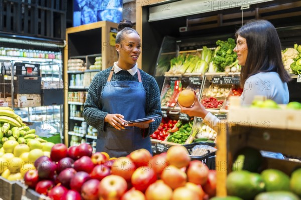 Supermarket employee assisting customer with grapefruit selection while checking prices and availability on a tablet among fresh organic products