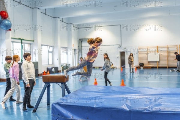 Elementary school girl practicing gymnastics, jumping over a pommel horse in the school gym watched by her classmates and teacher