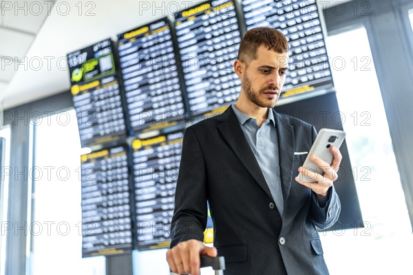 Businessman holding suitcase and using smartphone while checking flight schedule information on airport timetable display