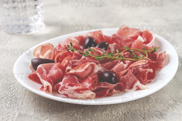 Freshly cut dried meat, served with olives and herbs, on a white plate, light background, no people
