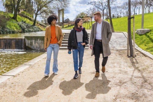 Three young professionals are enjoying a walk and conversation together in a sunny park, strolling along a path by a pond