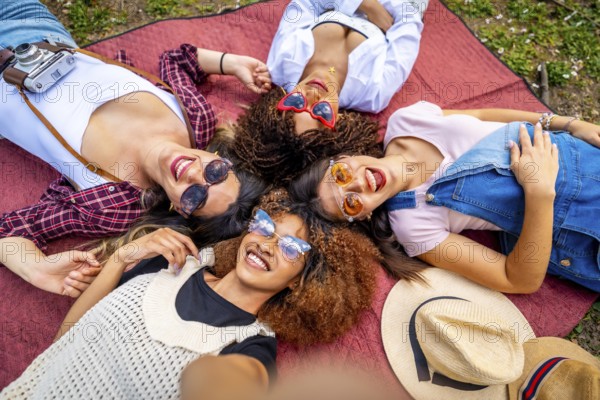 Four cheerful young women with sunglasses and hats are lying in circle on a blanket at a park, taking a selfie