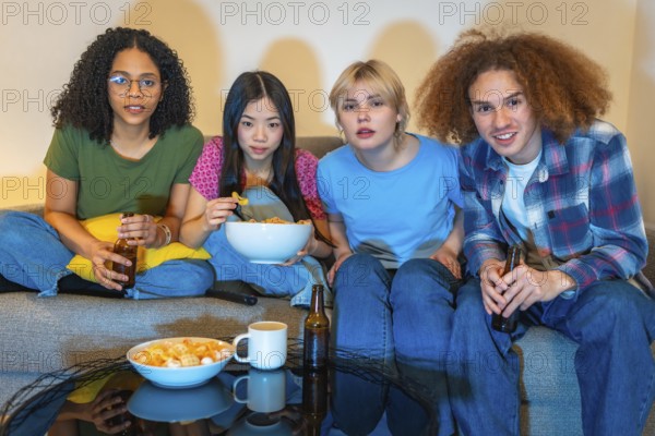 Four young friends enjoying a night in, watching television, eating snacks and drinking beer, sitting comfortably on a sofa