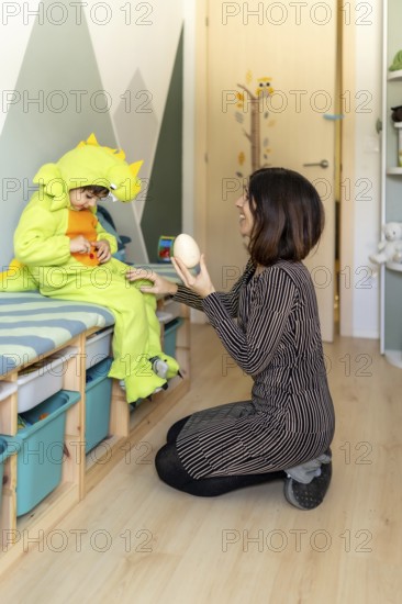 Mother kneeling and showing a dinosaur egg to her son wearing a dinosaur costume while playing in his bedroom