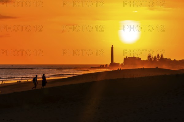 Tourists walking on the maspalomas dunes at sunset, with the maspalomas lighthouse in the background, gran canaria, canary islands, spain