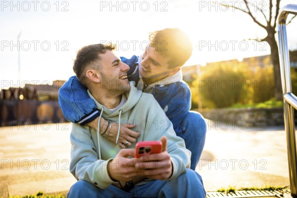 Two men hugging and laughing while using a smartphone outdoors, enjoying a sunny day together