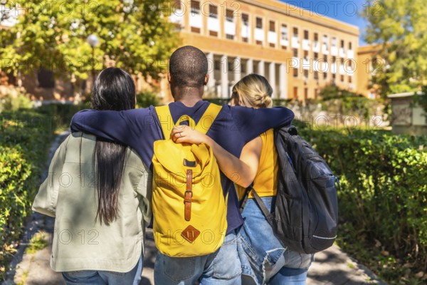 Rear view of three multi-ethnic students embracing while walking together to the university building