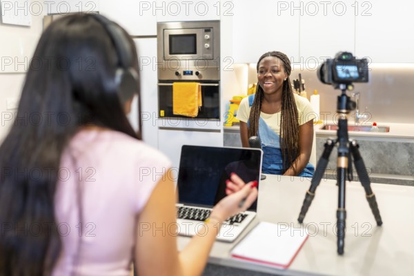 Two teenage girls creating content for their video blog, recording with a camera and using a laptop in a home kitchen