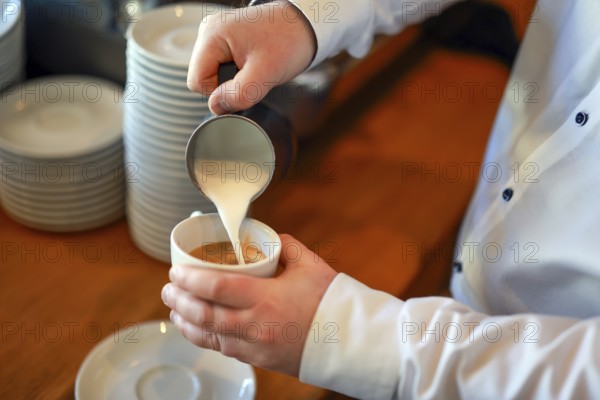Germany - Barista prepares a coffee, here a cappuccino, hand pours milk foam from a pot into a cup with espresso coffee