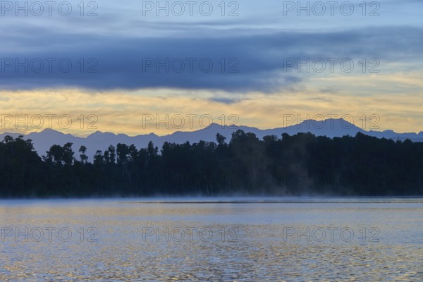 A hazy lake at sunrise with a mountainous silhouette and blue surroundings, Lake Mahinapua, Ruatapu, South Island, New Zealand
