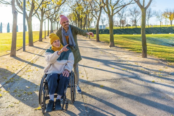 Two friends are walking in a park, one is using a wheelchair and they are both pointing at something interesting in the distance