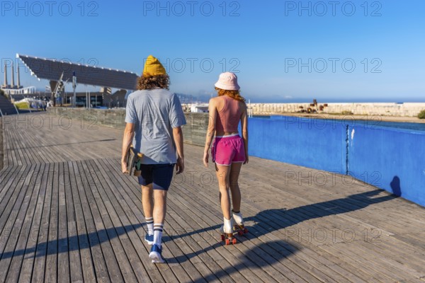 Young man carrying a skateboard strolls alongside a roller skating woman on a wooden pier, enjoying a sunny day by the sea