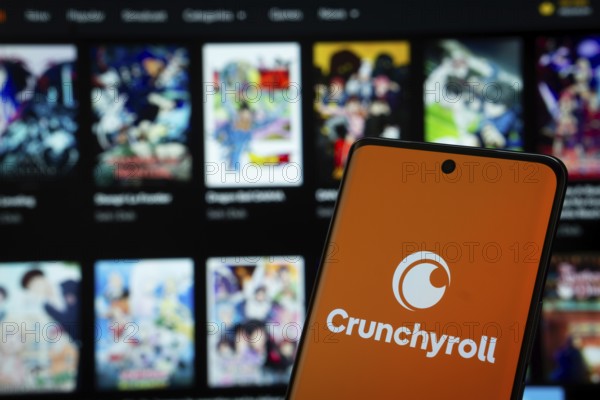 Crunchyroll logo is displayed on a smartphone screen, with the crunchyroll website visible on a computer screen in the background
