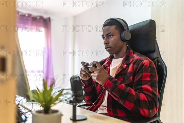 Young man playing videogames on a desktop computer using a controller, wearing headphones and using a microphone