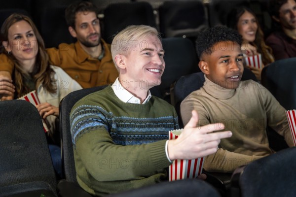 Group of diverse friends watching a film at the movie theater, commenting the scene and smiling, while eating popcorn