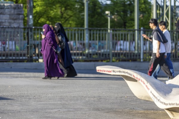 New seating on Norbert-Burger-Platz at the end of the Hohenzollern Bridge on the right bank of the Rhine. Inscription in various languages: What do you want in Cologne? Three Muslim women with burqas. Cologne, North Rhine-Westphalia, Germany