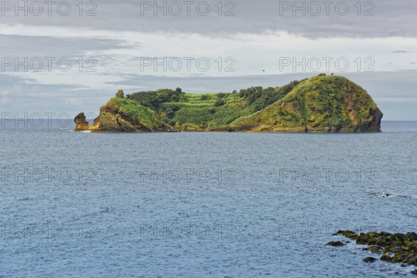 Green, rocky island and protected area Ilhéu de Vila Franca do Campoin a calm sea under a cloudy sky, Vila Franca do Campo, Sao Miguel, Azores, Portugal